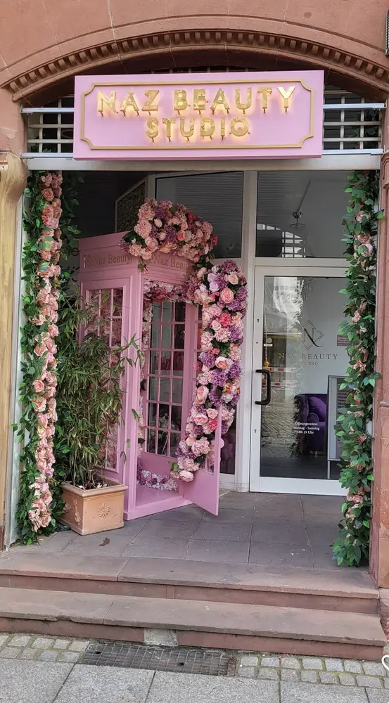 Eingang des Naz Beauty Studio in Durlach: rosa Schild mit goldenen Buchstaben, Blumengirlanden am Portal und davor die rosa Telefonzelle voller Blumen